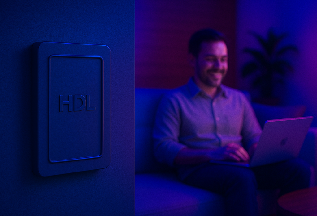 A modern HDL smart home switch is mounted on a blue wall, while in the background, a man relaxes on a couch with a laptop in a room with ambient purple and blue lighting